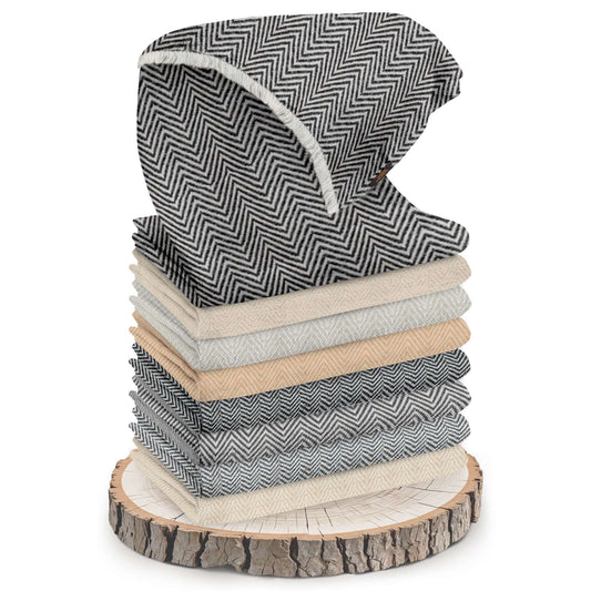 Das Bild zeigt einen Stapel mehrerer gefalteter Schals oder Tücher, die übereinander auf einer runden Holzscheibe liegen. Die Schals haben unterschiedliche Grautöne und Beige-Nuancen mit mittlerem Fischgrätenmuster. Das oberste Tuch ist teilweise aufgeschlagen, sodass das Muster deutlich zu sehen ist und an der Kante sind feine Fransen erkennbar. Die Holzscheibe darunter dient als dekorative Unterlage und hat eine sichtbare Baumrinde.